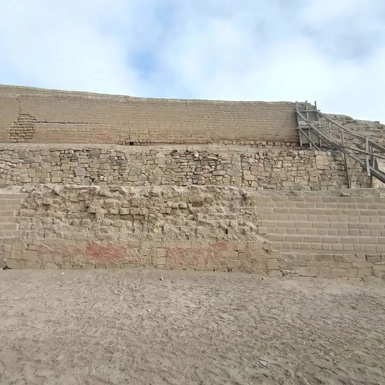Terraced adobe and stone walls at Pachacamac retain patches of reddish plaster, with a wooden staircase on the right.