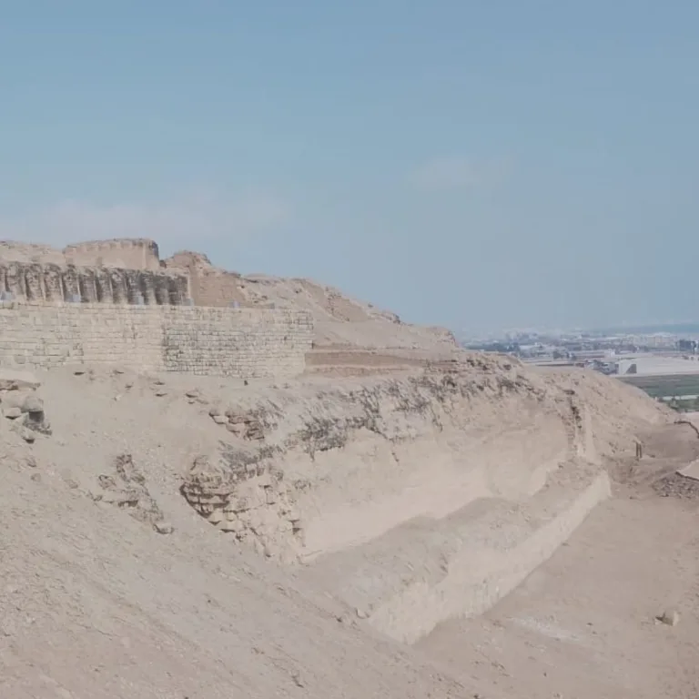 Eroded terraces and exposed masonry at Pachacamac overlook the coastal plain beneath a hazy blue sky.